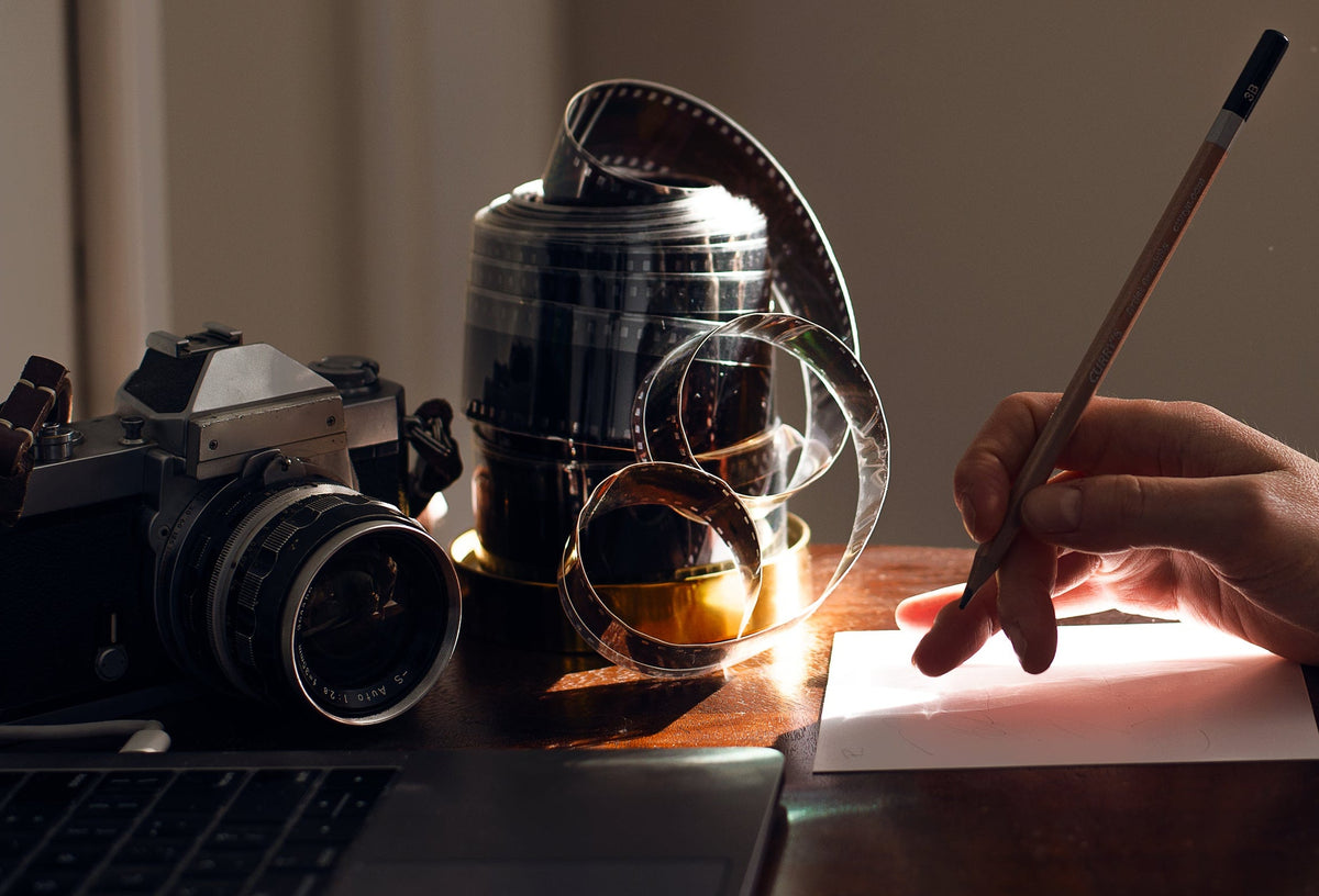 Person writing with a pen on paper next to a camera and film reels on a desk.
